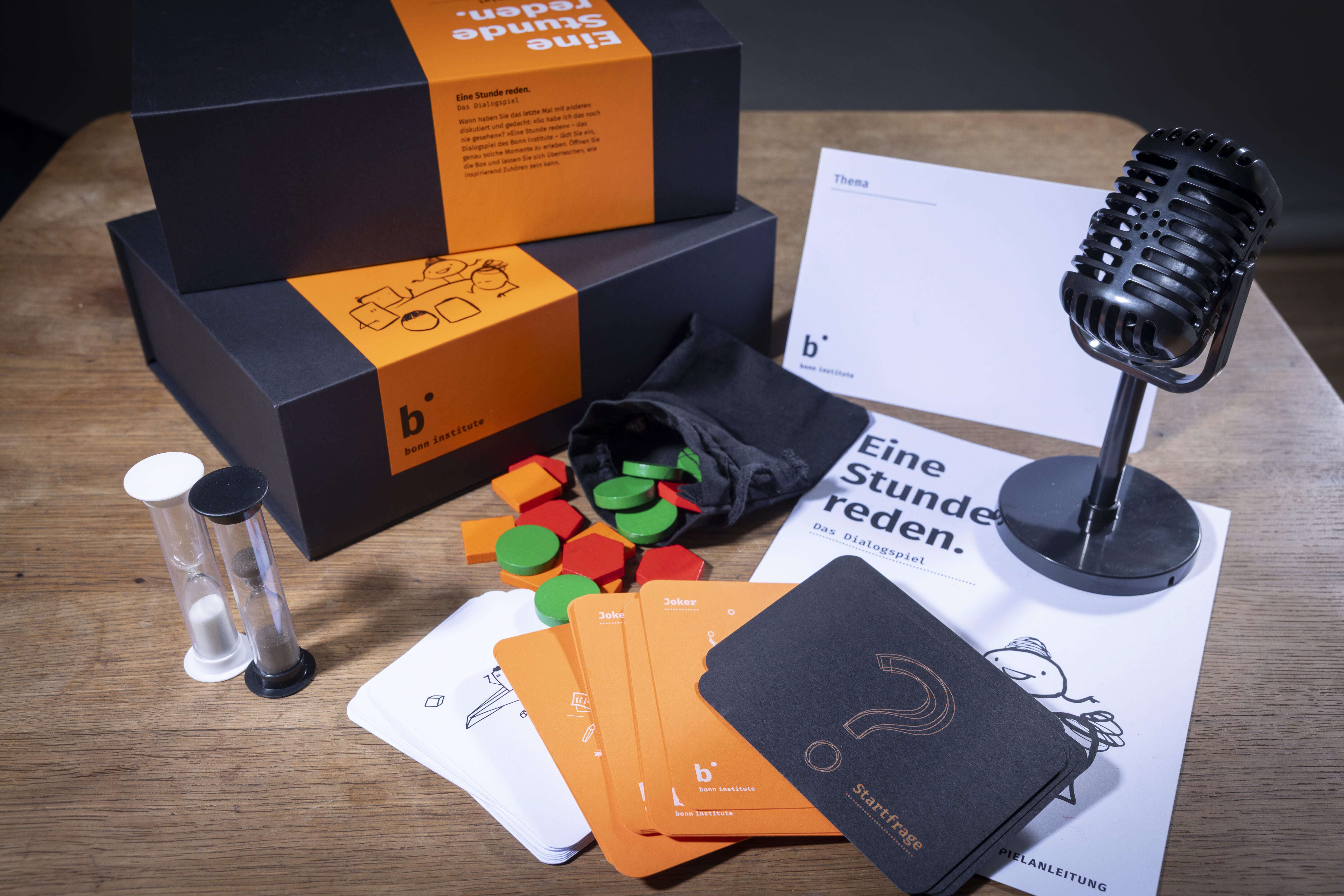 Das Spielset von "Eine Stunde reden" liegt ausgebreitet auf einem Tisch. Zu sehen sind der Spielkarton, eine Sanduhr, ein Mikrofon, eine Spielanleitung und verschiedene Spielteile- und karten.