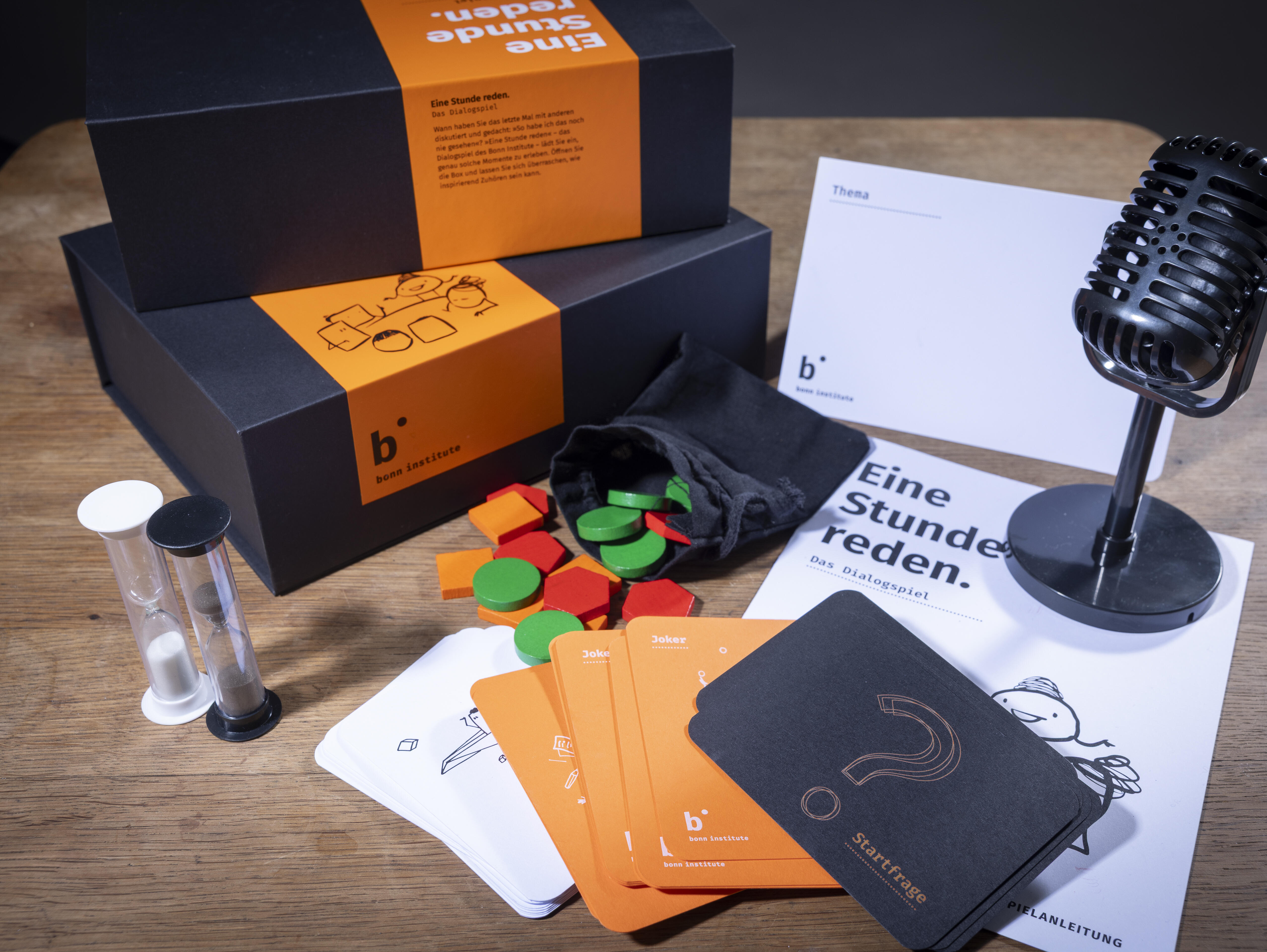 Das Spielset von "Eine Stunde reden" liegt ausgebreitet auf einem Tisch. Zu sehen sind der Spielkarton, eine Sanduhr, ein Mikrofon, eine Spielanleitung und verschiedene Spielteile- und karten.