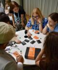 A group of people play the cooperative discussion board game One Hour, Many Voice at a round table