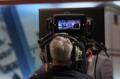 A news show cameraman sits behind the camera which is focused on a trio of figures in a news program,