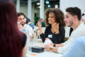 drei Mitspielende sitzen am Tisch, die mittlere Person spricht rege, die anderen beiden hören zu