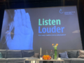 Screen mit Logo der Konferenz "Listen Louder", davor Bühnensetting.
