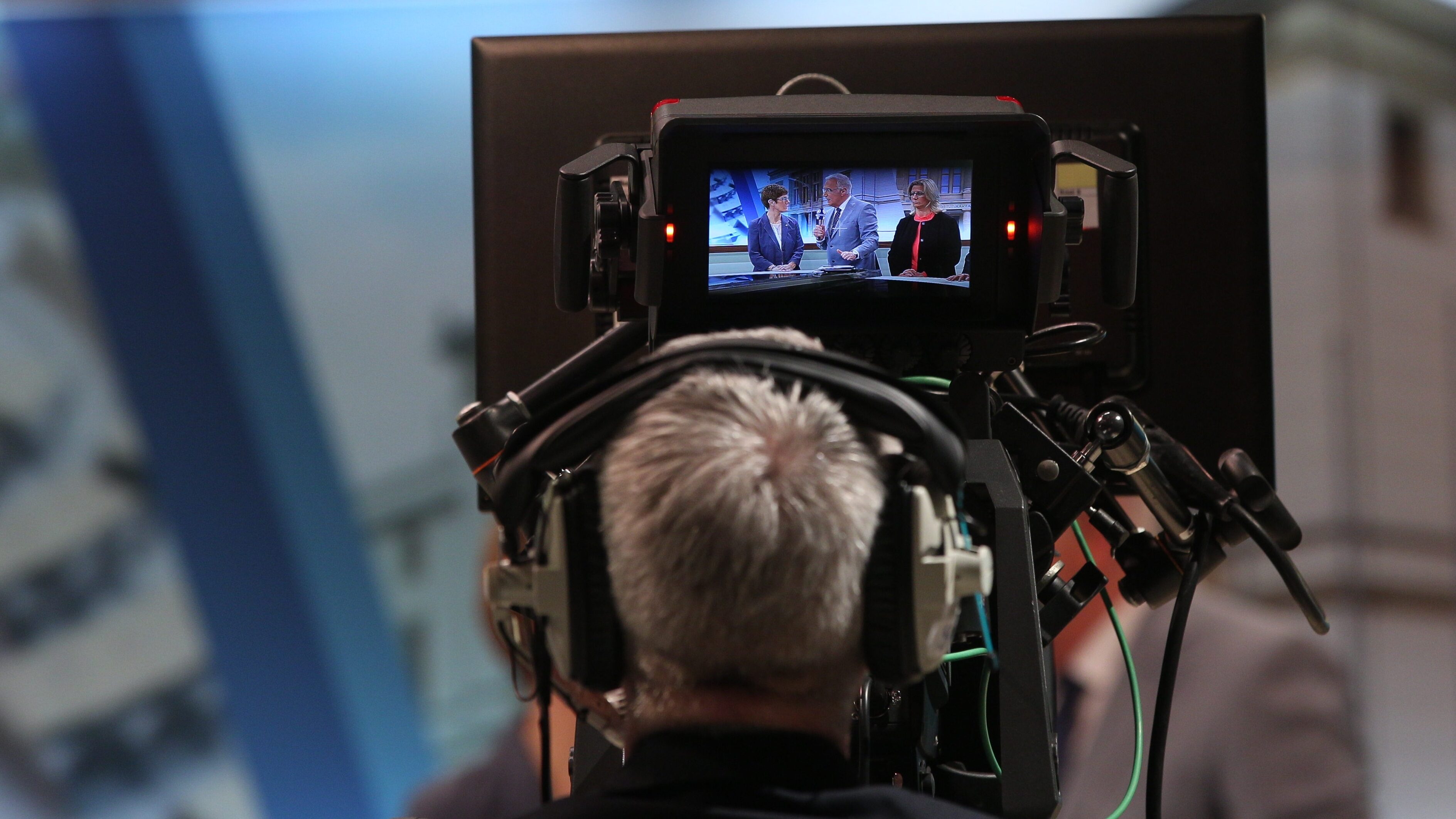 A news show cameraman sits behind the camera which is focused on a trio of figures in a news program,