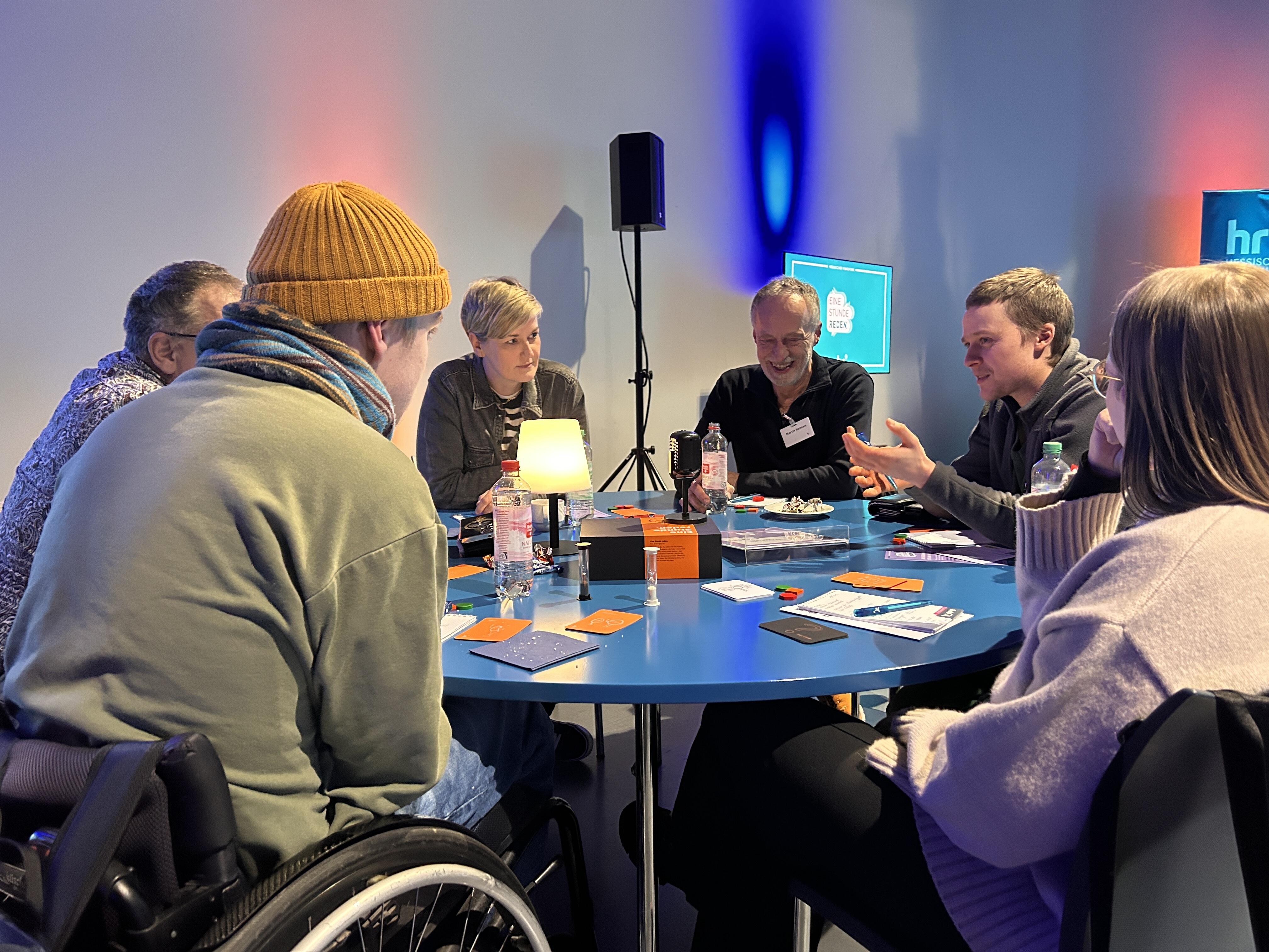 Eine Gruppe von Menschen sitzt an einem runden Tisch und spielt "Eine Stunde reden. Das Dialogspiel"