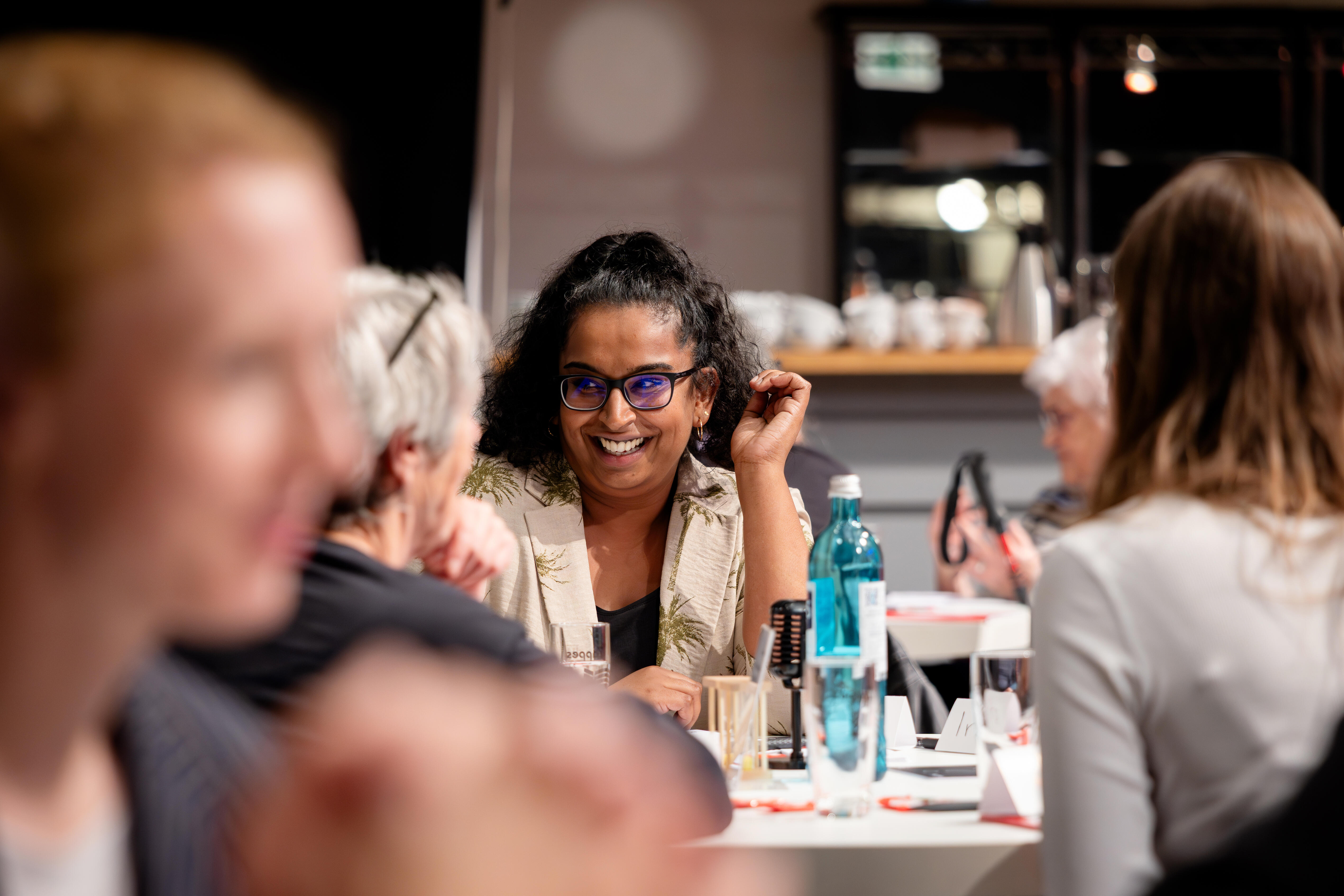 Im Bild zu sehen ist Kanimoli Pavanandarajah von der Sparkasse KölnBonn, sie leitet die Spielrunde an einem runden Tisch und lächelt die anderen Teilnehmenden an.