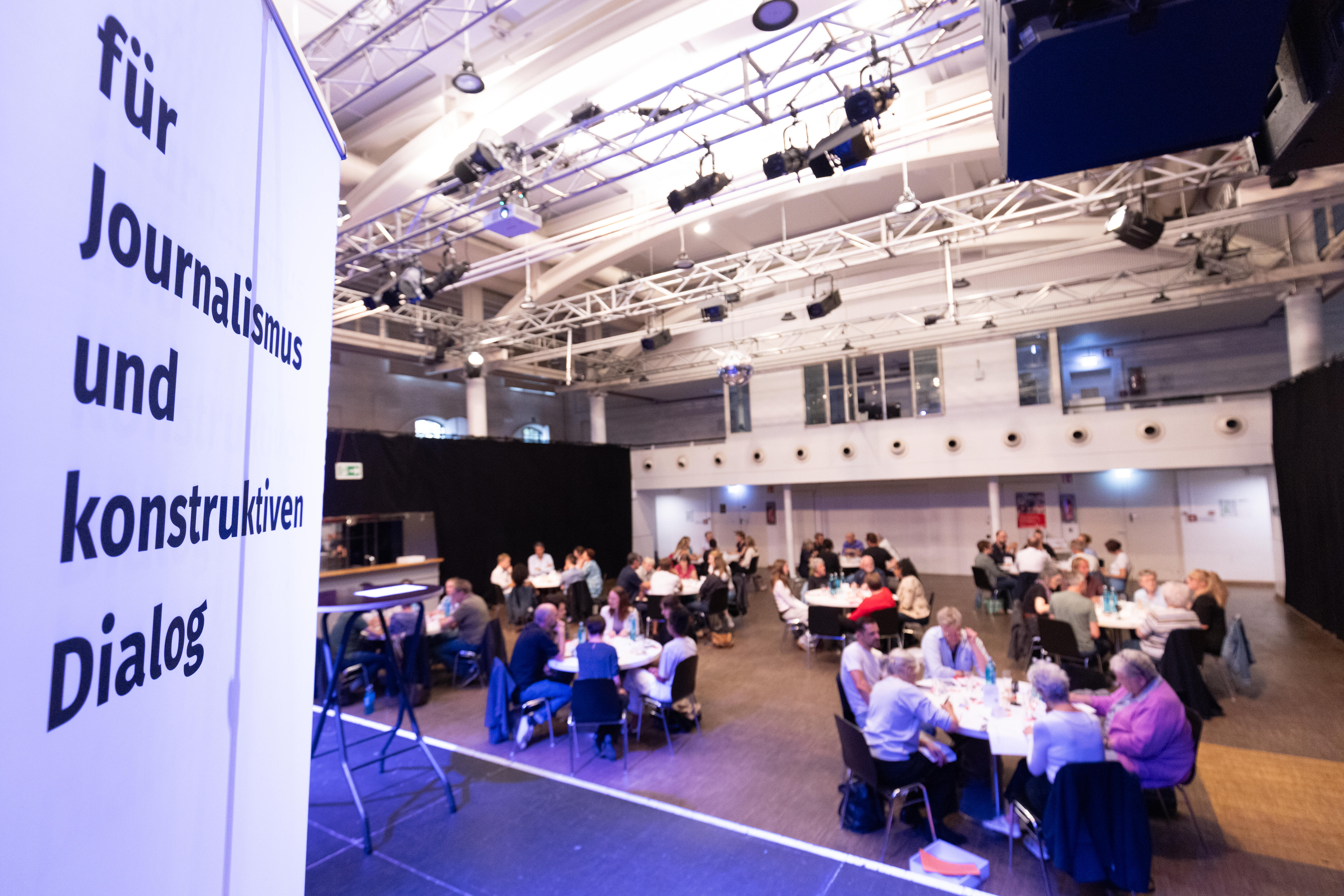 Links ein Banner auf dem der Slogan des Bonn Institute zu lesen ist: "Für Journalismus und konstruktiven Dialog". Rechts sitzen Menschen an runden Tischen und spielen "Eine Stunde reden".