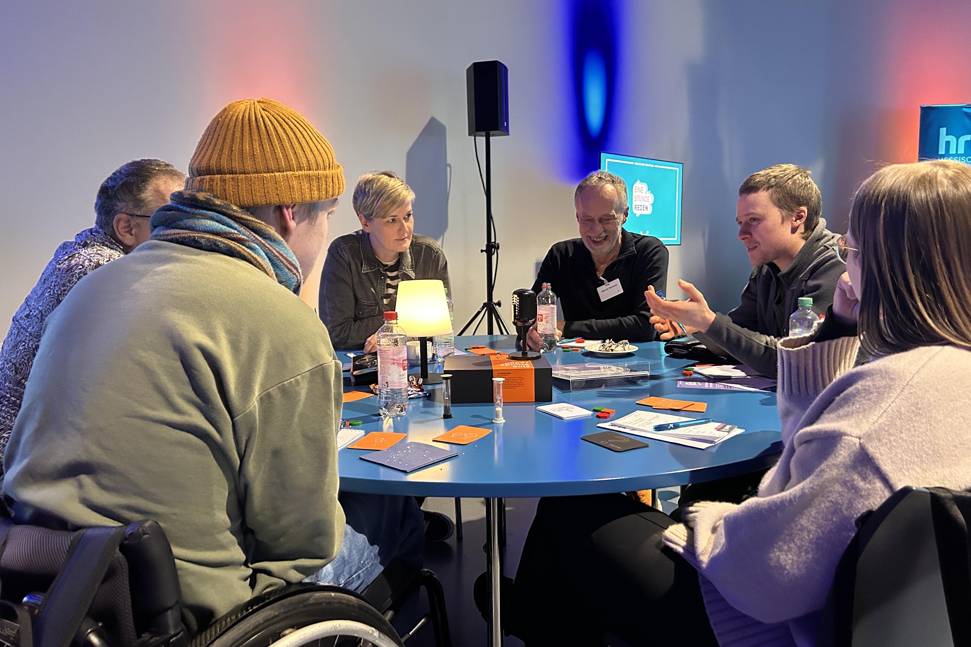 Sechs Menschen sitzen an einem runden Tisch und spielen "Eine Stunde reden" in Kassel.